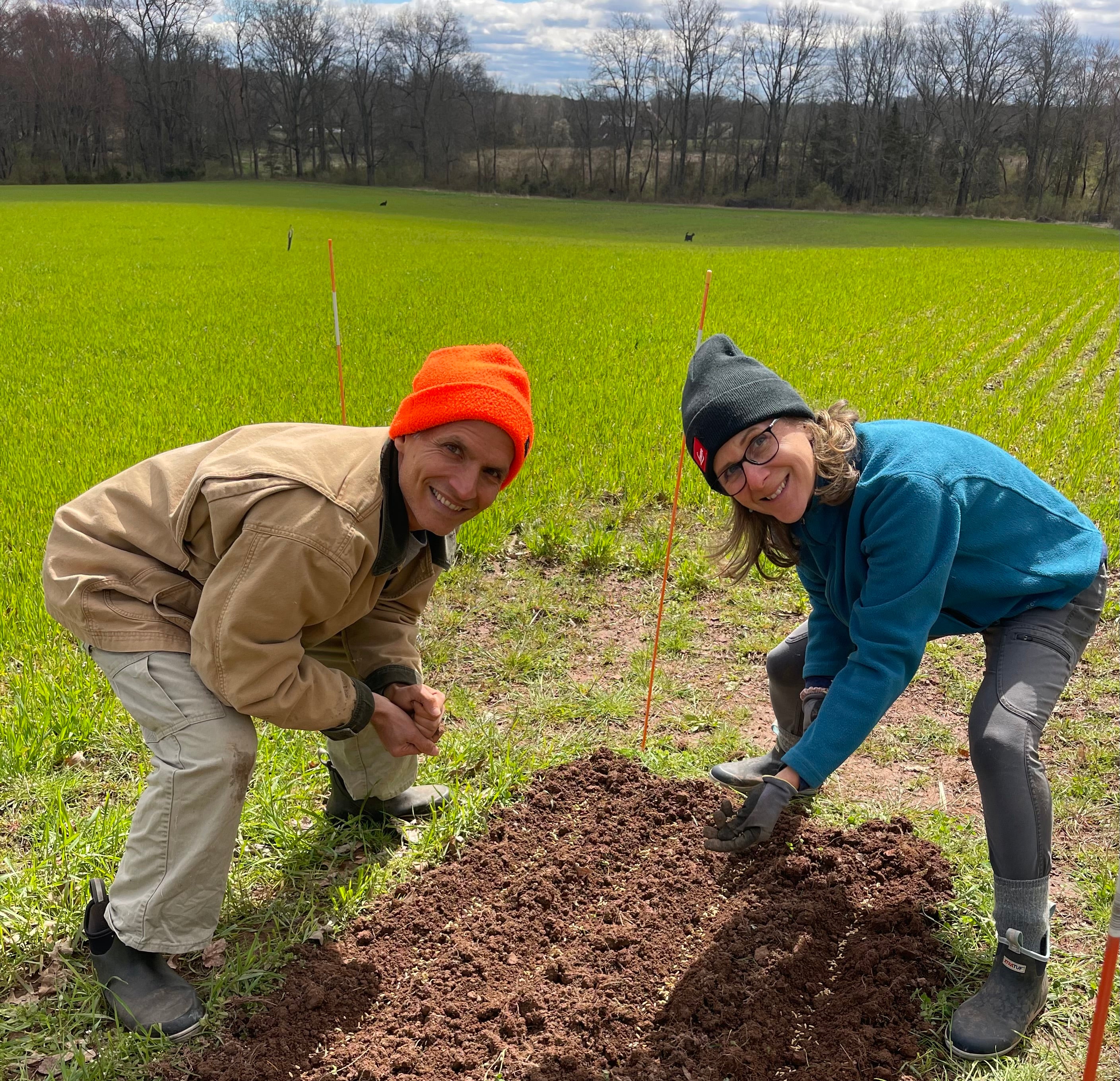 Two people crouching in a green field next to freshly turned soil, wearing outdoor clothing including an orange beanie, tan jacket, blue fleece, and dark beanie.
