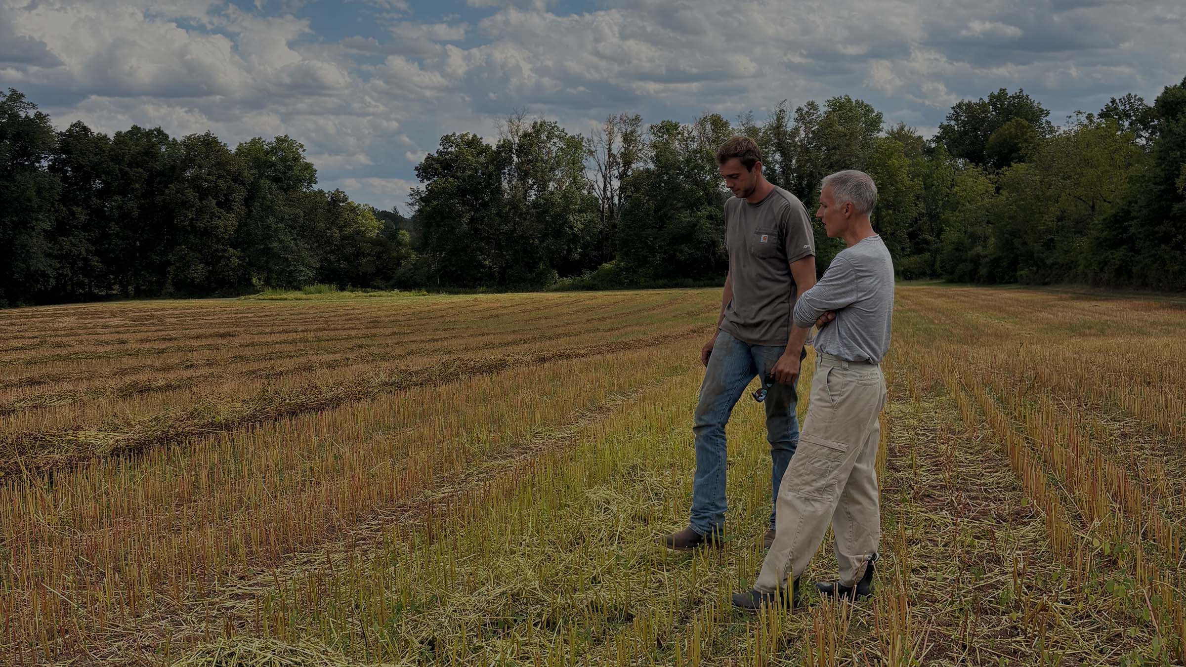 Two men standing in a harvested field under a cloudy blue sky, with trees in the background.