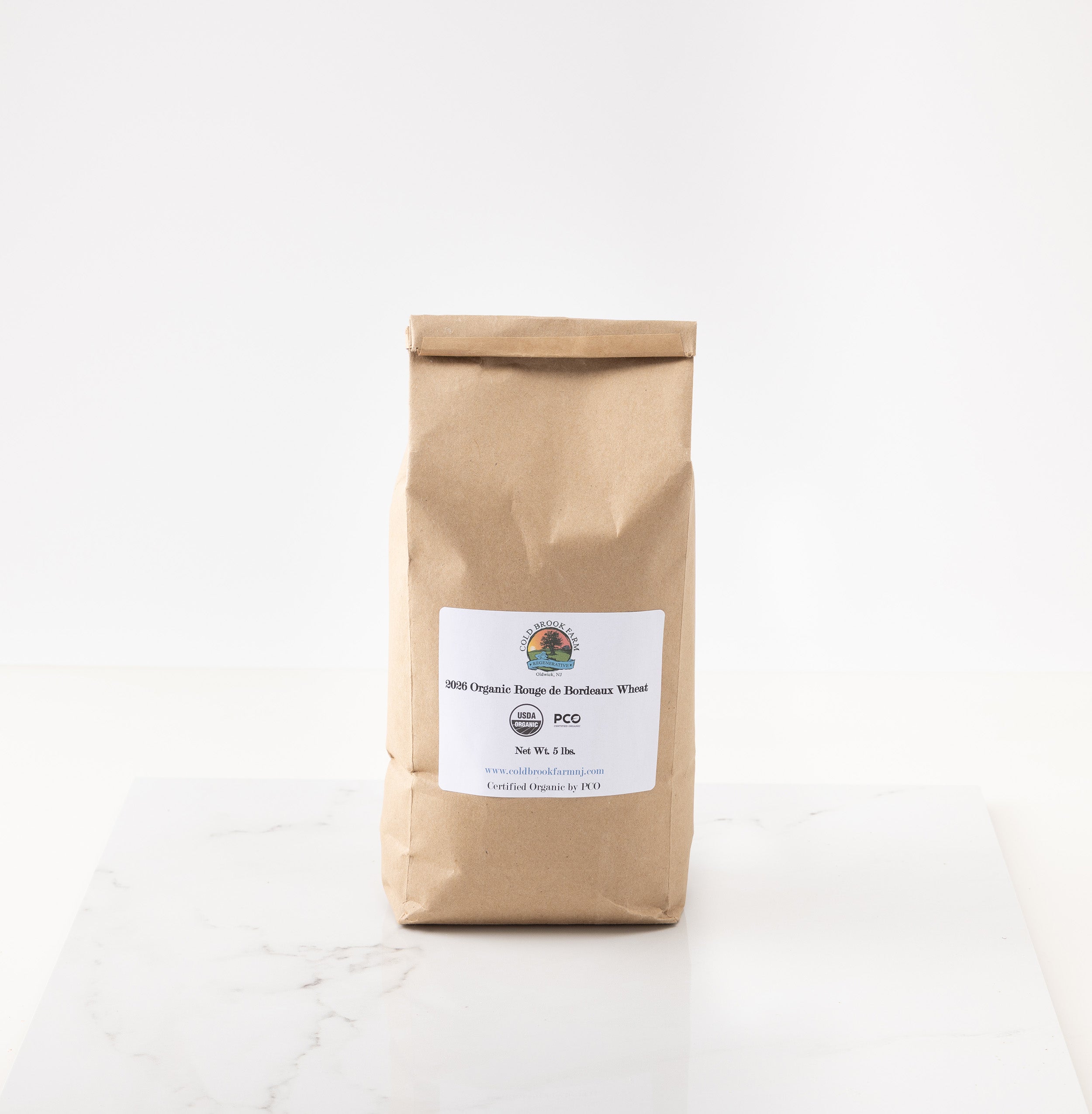 A brown kraft paper bag of rouge de bourdeaux wheat standing upright on a white marble surface against a white background.