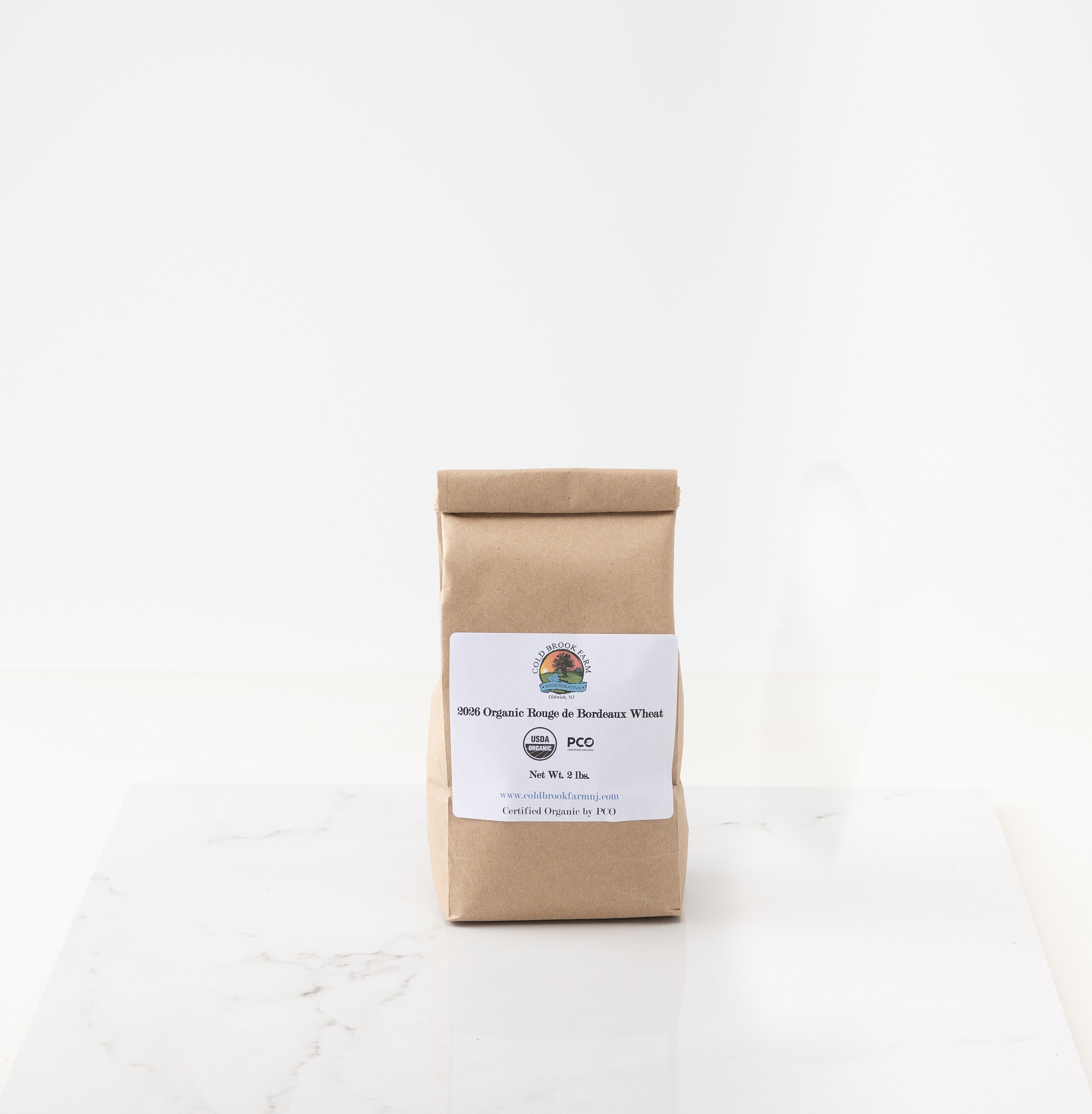 A kraft paper bag of organic rouge de bourdeaux wheat, featuring a white label with USDA Organic and PCO logos, set on a white marble surface against a white background.