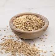A small round wooden bowl filled with grain oats porridge, with some loose oats scattered on a light marble surface.