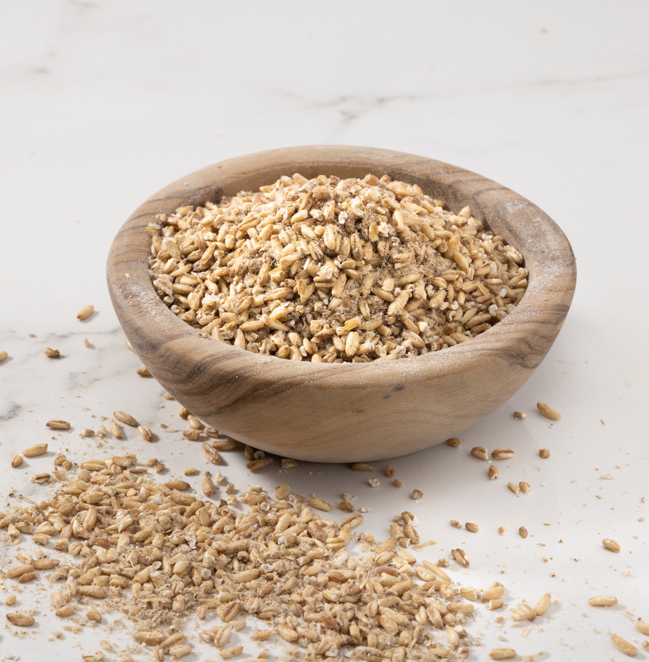 A small round wooden bowl filled with grain oats porridge, with some loose oats scattered on a light marble surface.