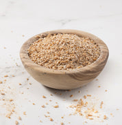 A small round wooden bowl filled with coarse ground Rouge de Bourdaux grains photographed on a light marble-like surface with a few grains scattered around.