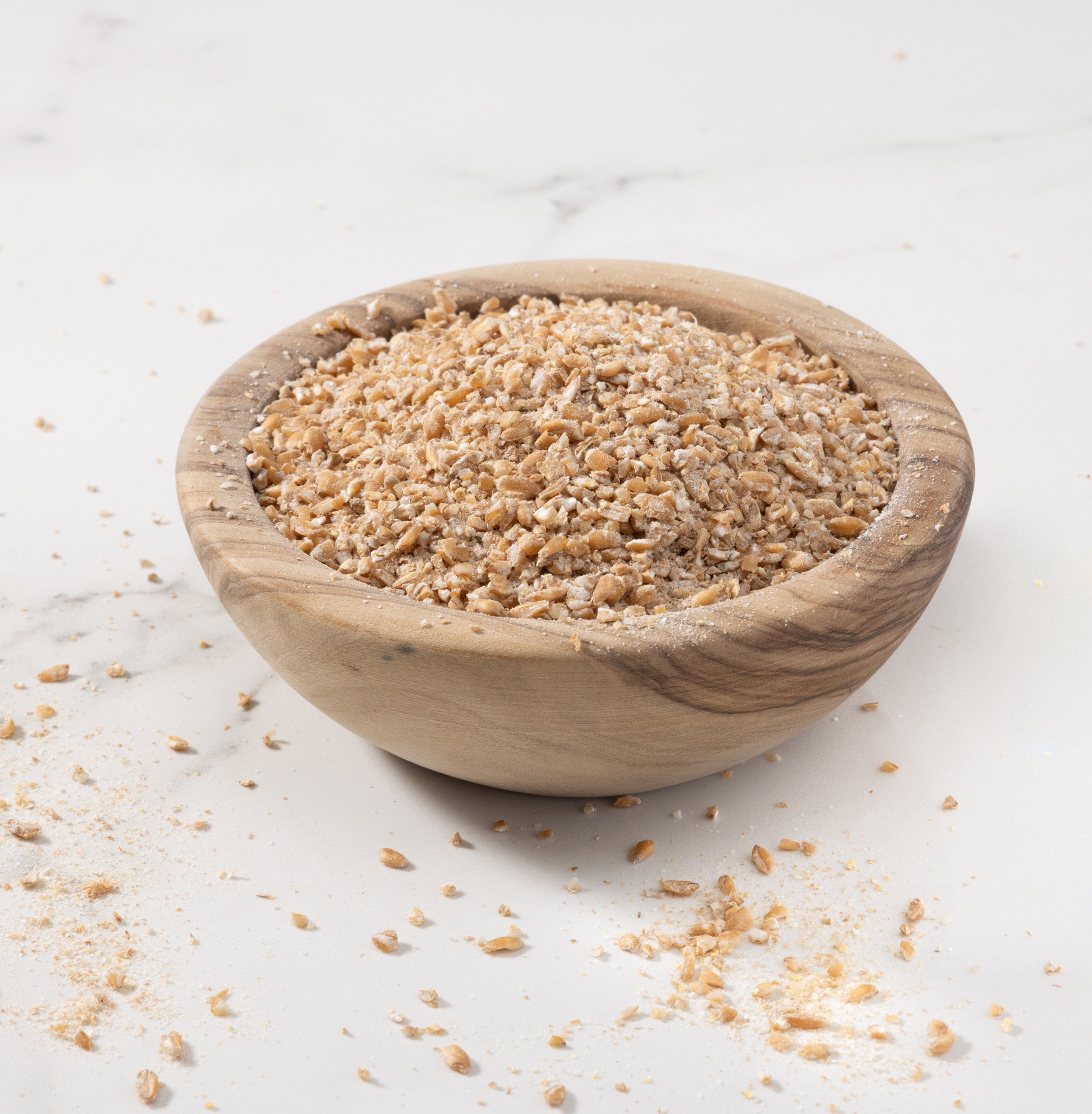 A small round wooden bowl filled with coarse ground Rouge de Bourdaux grains photographed on a light marble-like surface with a few grains scattered around.