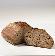 A rustic loaf of dark brown sourdough bread with one slice cut and placed in front, photographed on a clean white surface with a soft gray background.