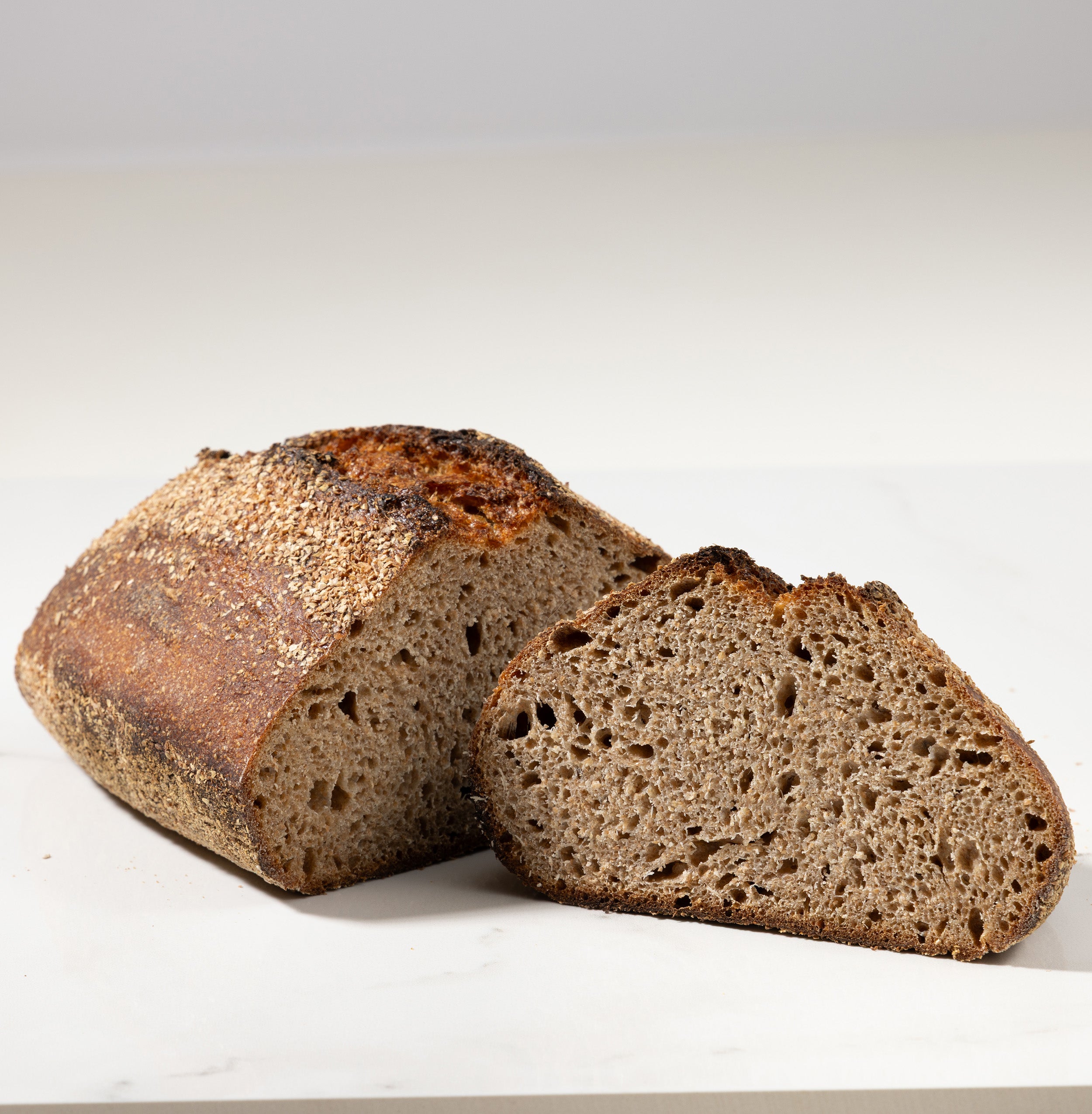 A rustic loaf of dark brown sourdough bread with one slice cut and placed in front, photographed on a clean white surface with a soft gray background.