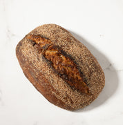A single rustic loaf of sourdough bread with a dark brown crust and a lighter baked crack, photographed from above on a white marble surface.