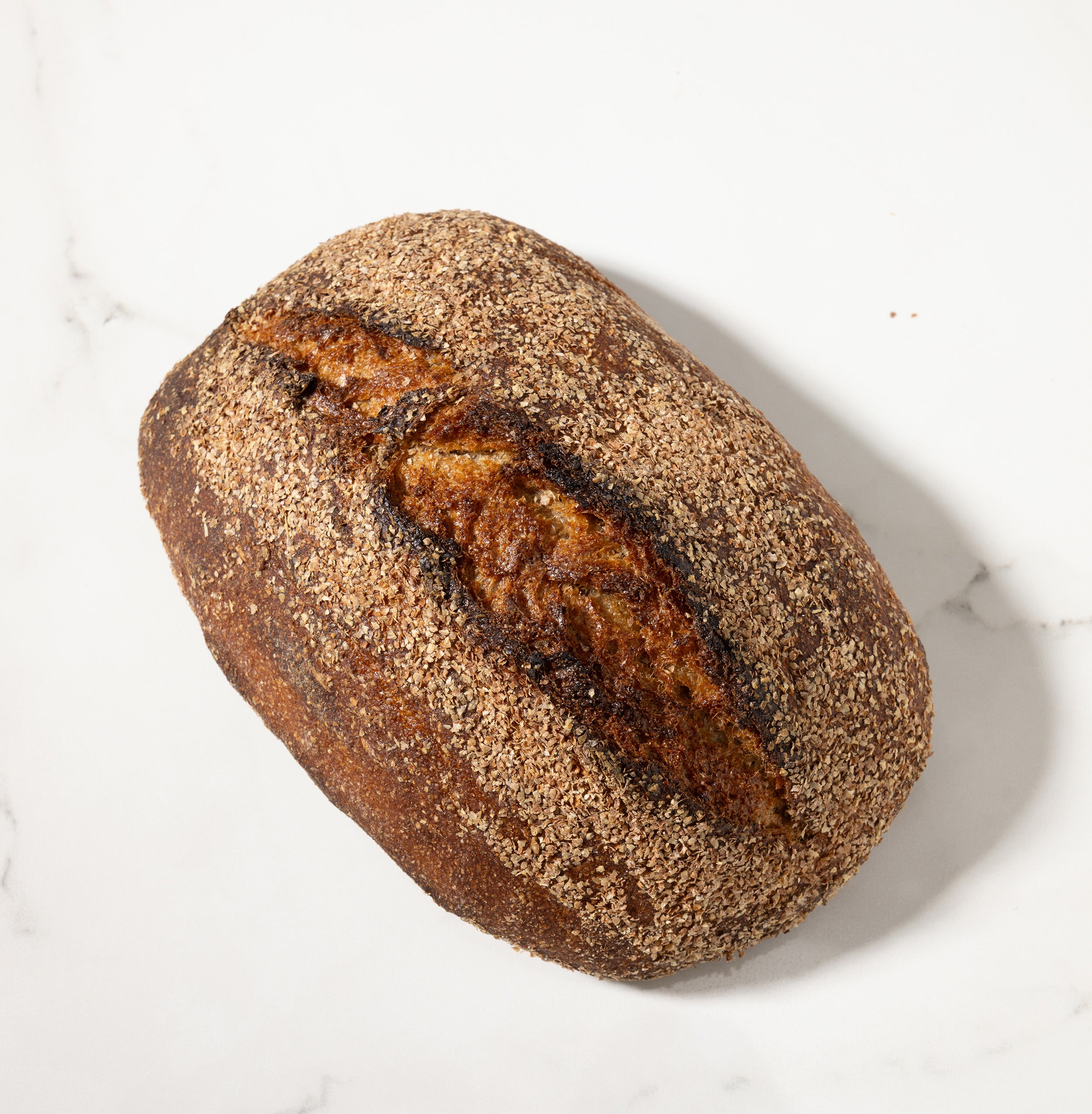 A single rustic loaf of sourdough bread with a dark brown crust and a lighter baked crack, photographed from above on a white marble surface.