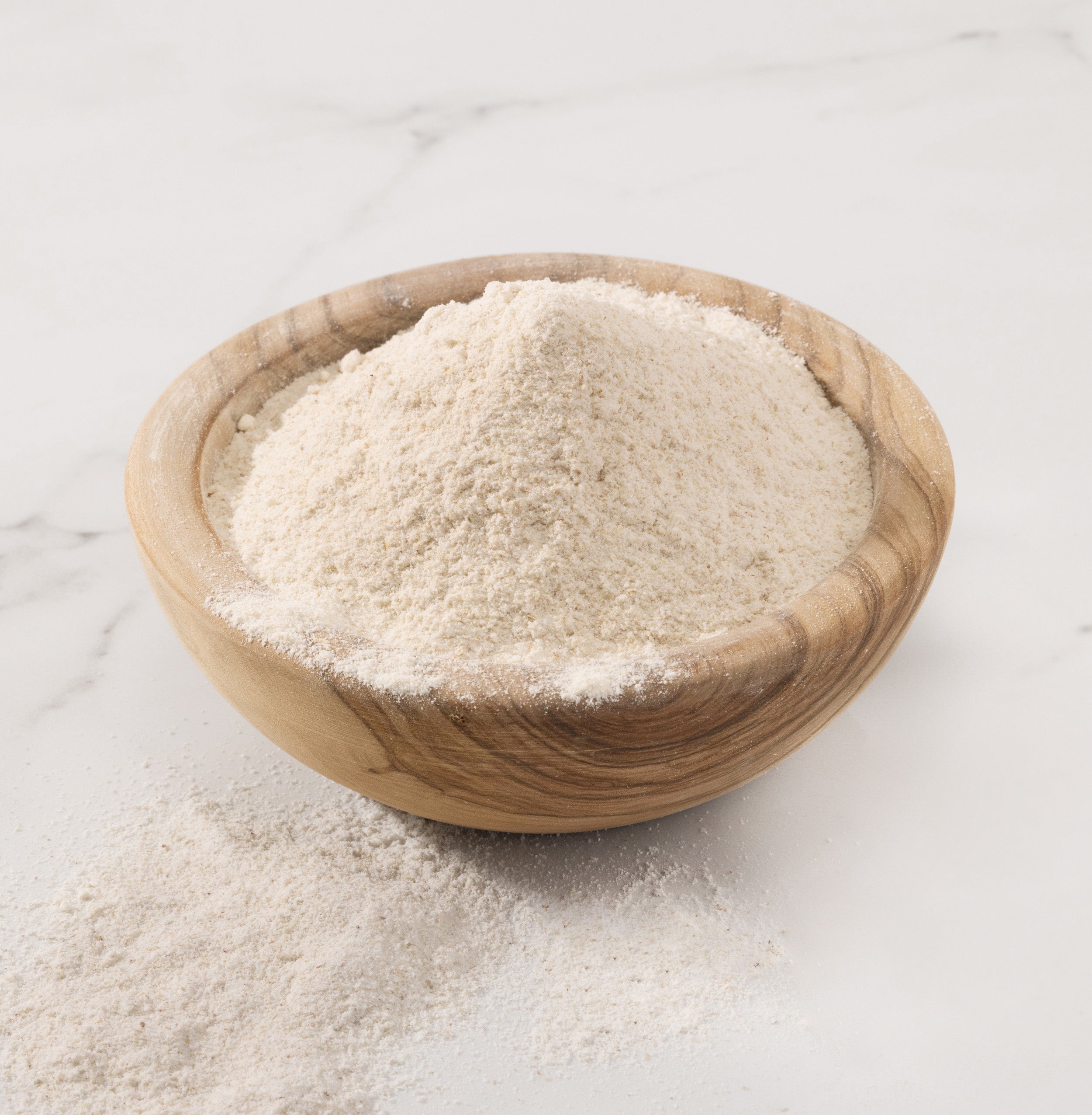 A mound of fine buckwheat flour, displayed in a small round wooden bowl on a light marble surface with some flour scattered outside the bowl.