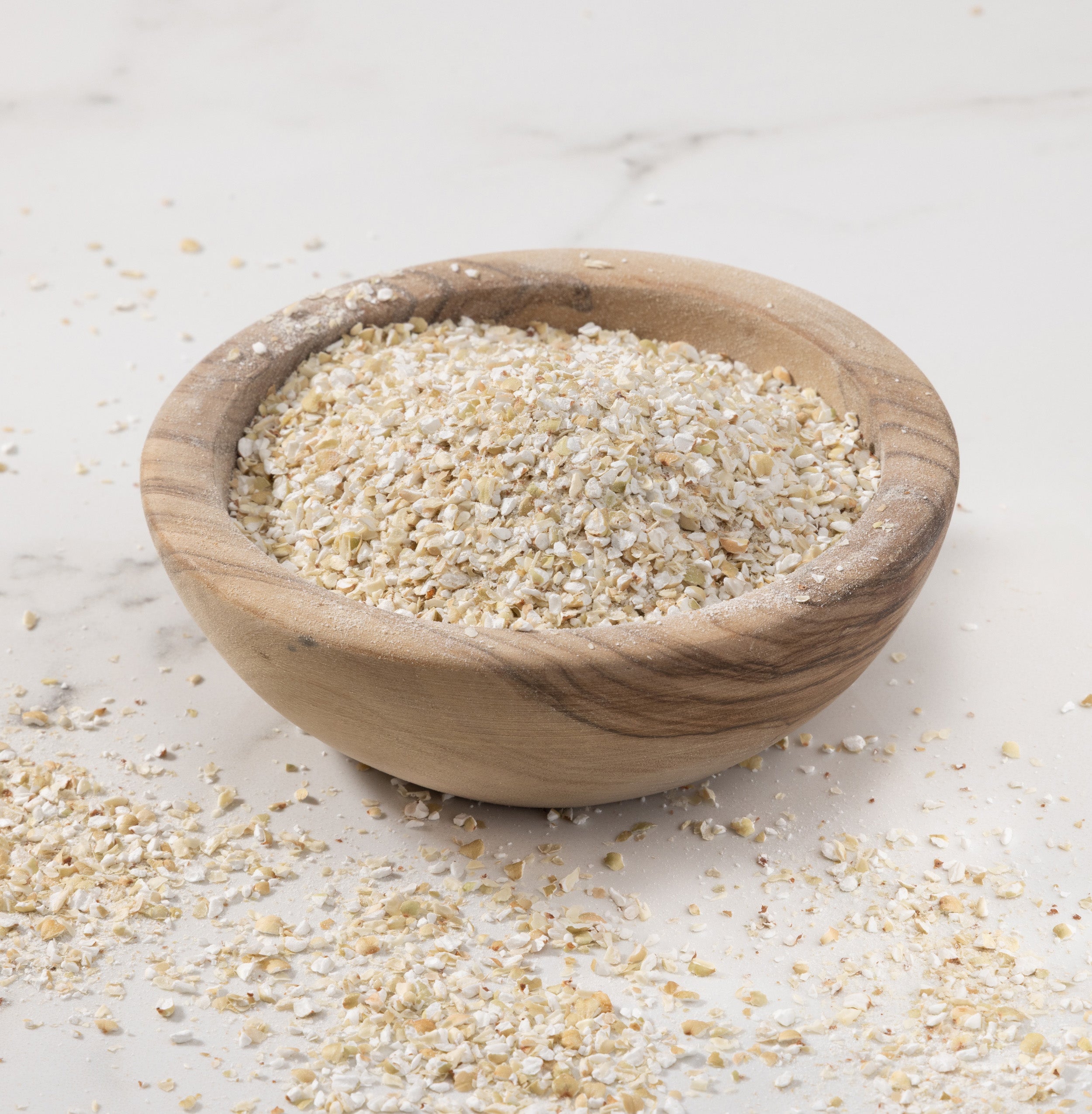 A small round wooden bowl filled with coarsely-ground buckweat groats, set on a light marble-like surface with some flakes scattered around.