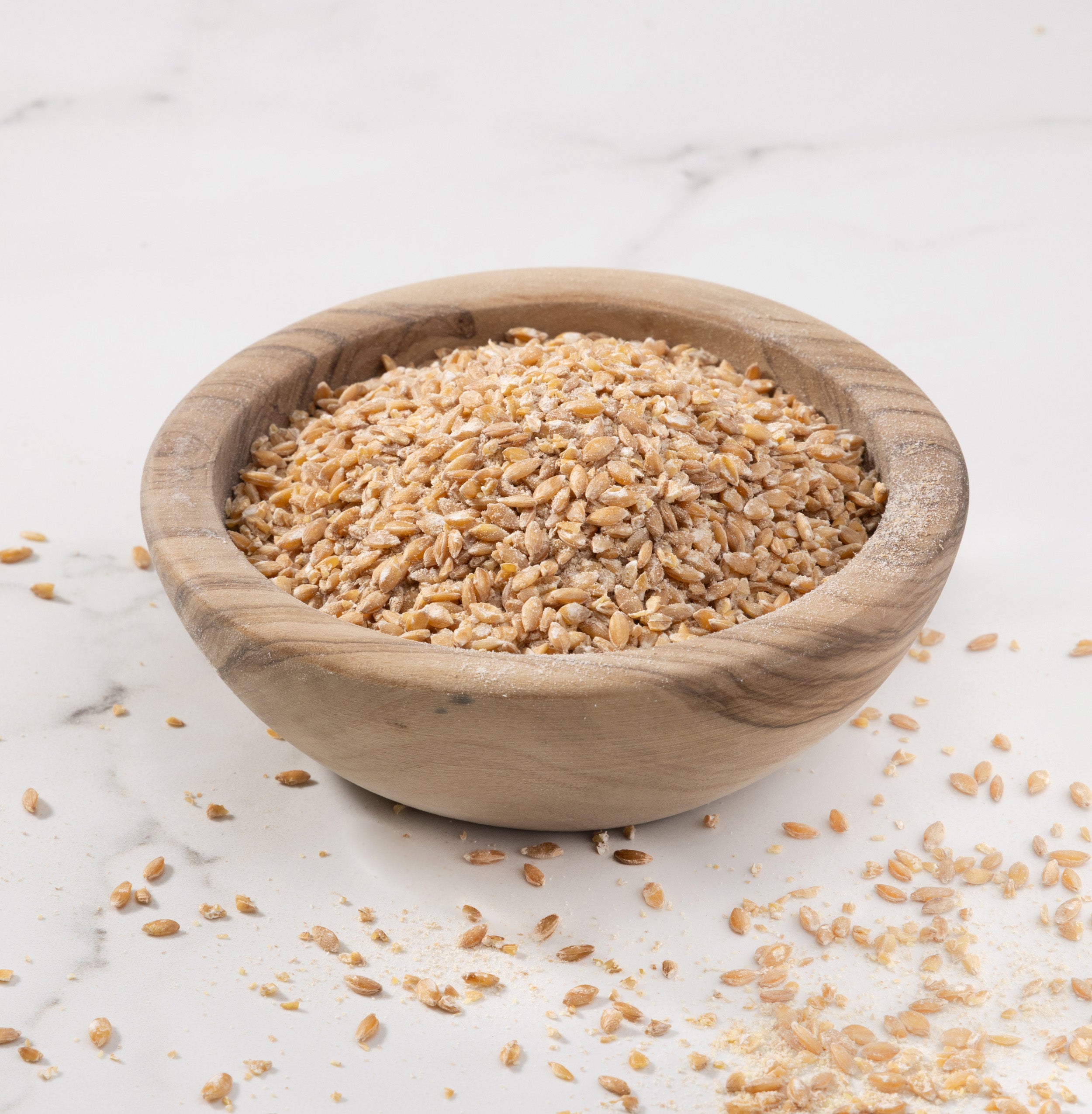 A small round wooden bowl filled with Einkorn porridge photographed on a light marble-like surface with a few grains scattered around.