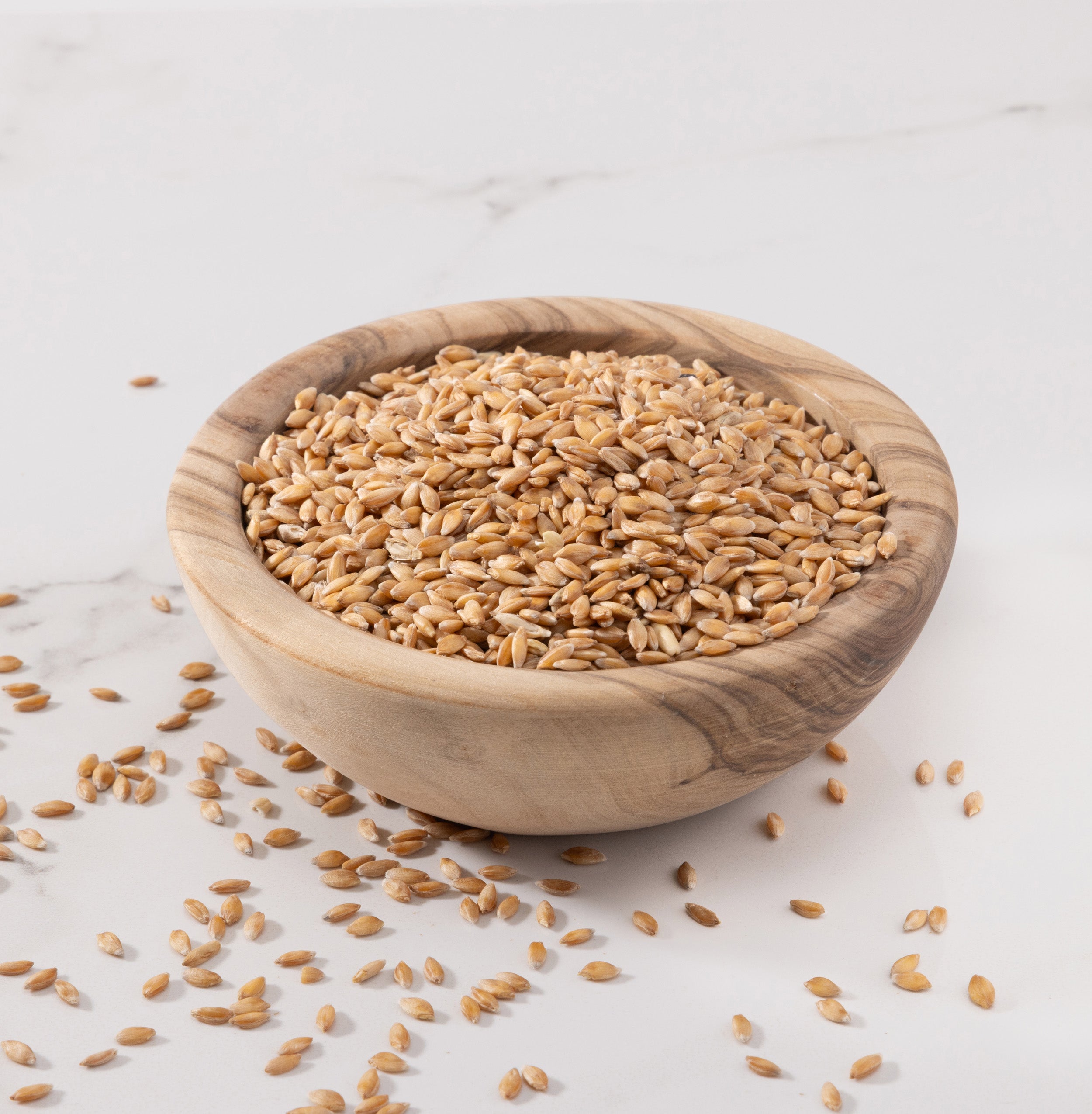 A small round wooden bowl filled with organic einkorn wheat berries, with a few grains scattered on a light marble surface.