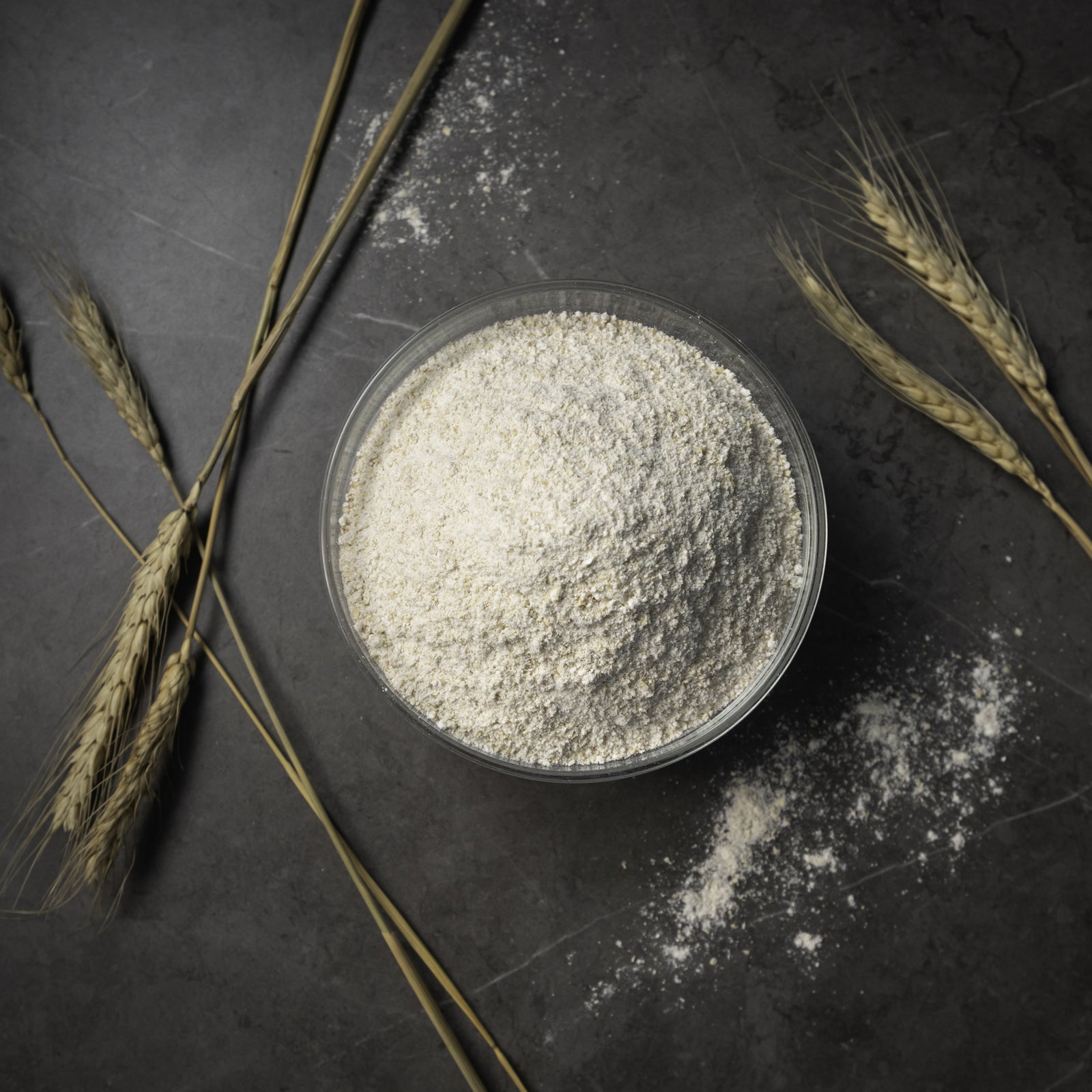 A clear round bowl filled with fresh-milled flour, arranged on a dark textured surface with scattered flour and wheat stalks around it.