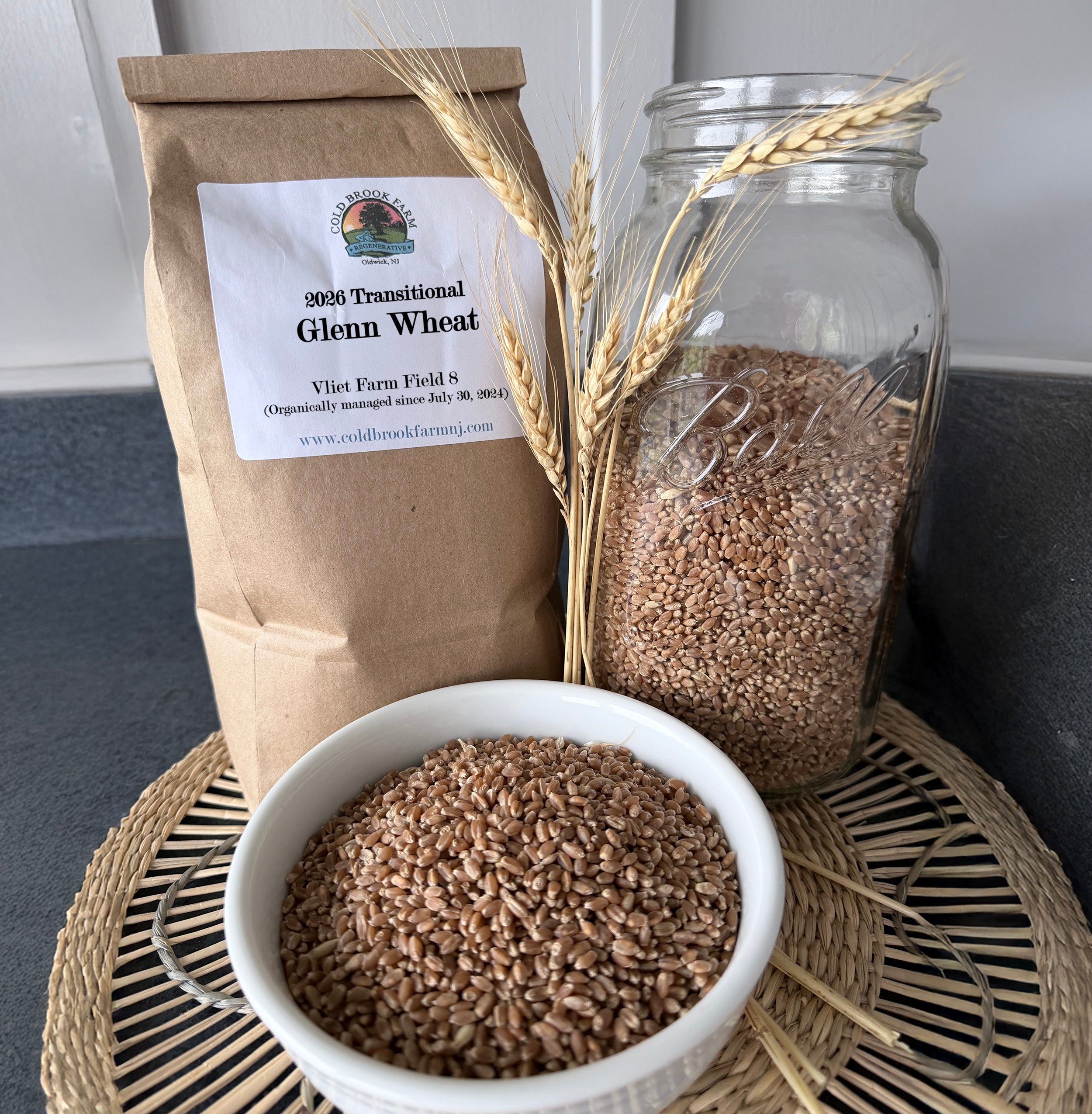 A kraft paper bag labeled 'Glenn Wheat' stands beside a clear glass jar filled with wheat grains and a white bowl of wheat, arranged on a woven tray with wheat stalks in a kitchen-like setting.