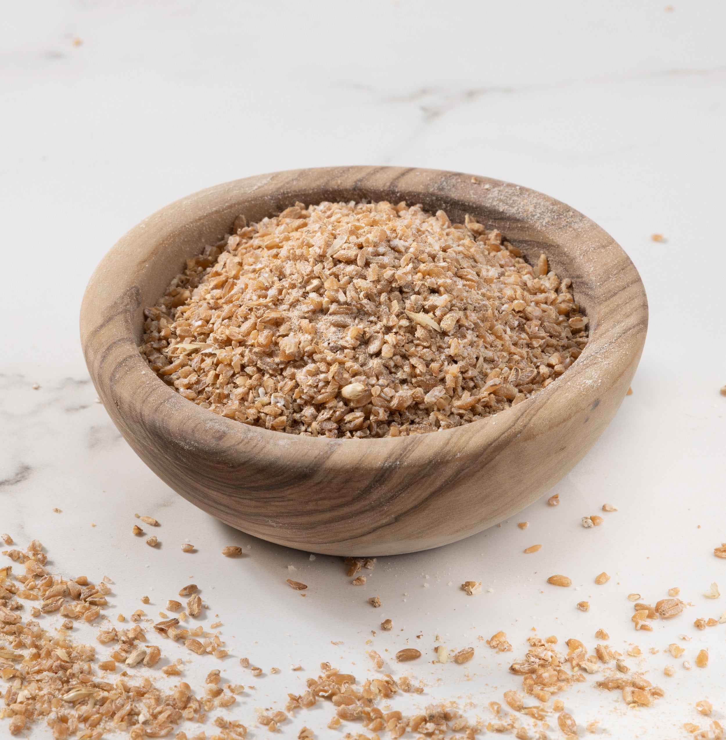 A small round wooden bowl filled with glenn wheat porridge photographed on a light marble-like surface with some crumbs scattered around.