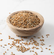 A wooden bowl filled with cold brook farm's baker's blend whole wheat grains, with a few scattered on a light marble surface.