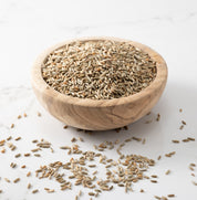 A wooden bowl filled with danko rye whole wheat grains, with a few scattered on a light marble surface.