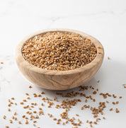 A wooden bowl filled with  whole glenn wheat grains, with a few scattered on a light marble surface.