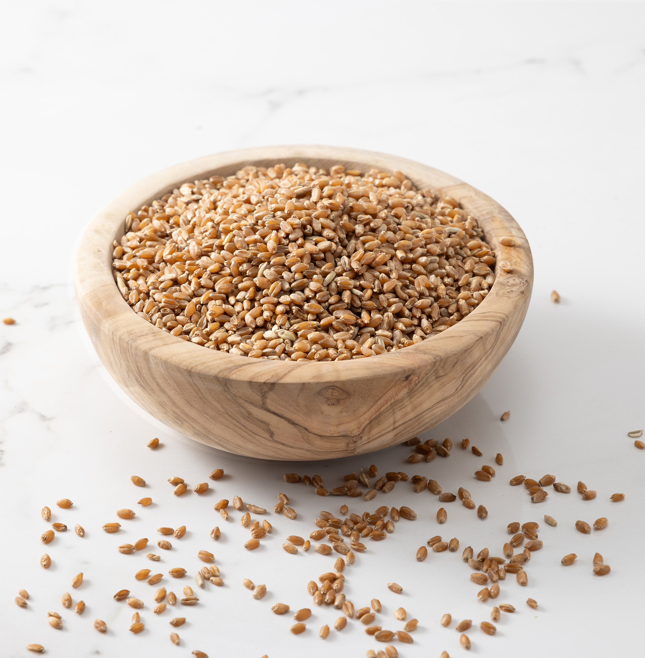 A wooden bowl filled with  whole glenn wheat grains, with a few scattered on a light marble surface.