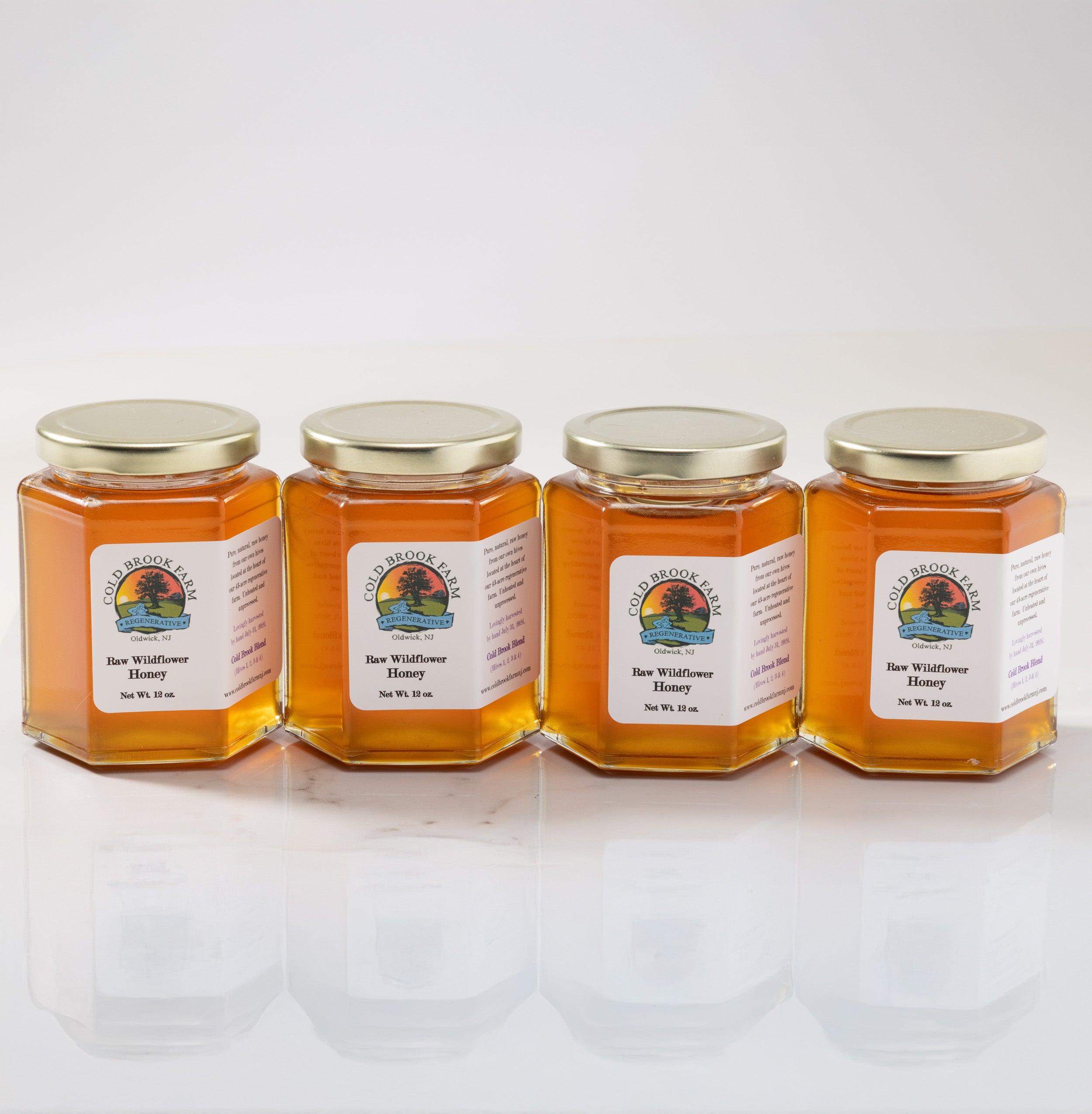 Four hexagonal glass jars of COLD BROOK FARM Wildflower Honey with gold lids and white labels, arranged in a row on a reflective white surface against a light gray background.