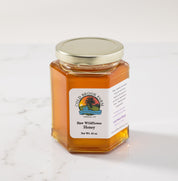 A hexagonal glass jar of Cold Brook Farm Raw Wildflower Honey with a white label and a silver metal lid, set on a white marble surface against a white background.