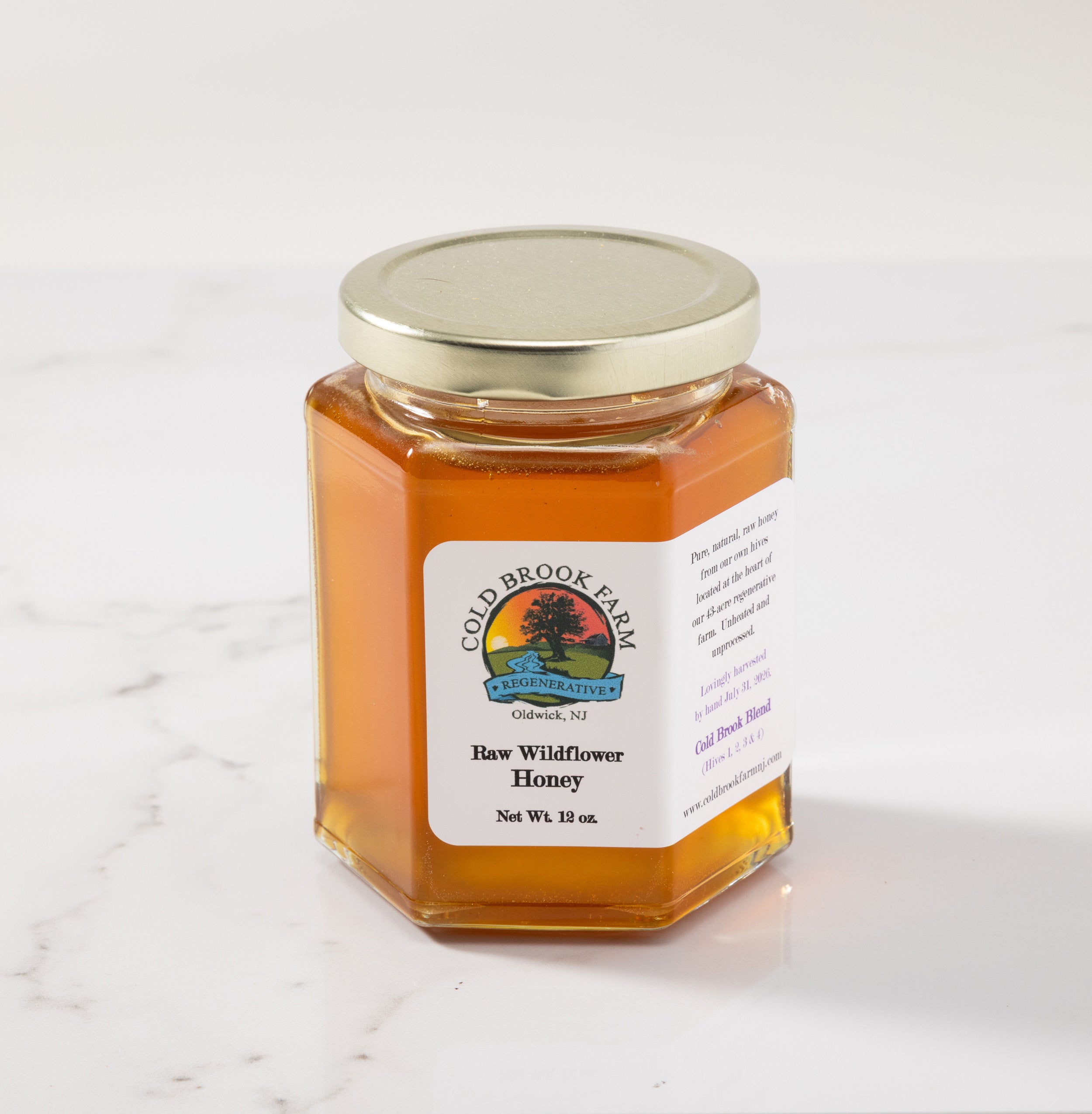A hexagonal glass jar of Cold Brook Farm Raw Wildflower Honey with a white label and a silver metal lid, set on a white marble surface against a white background.