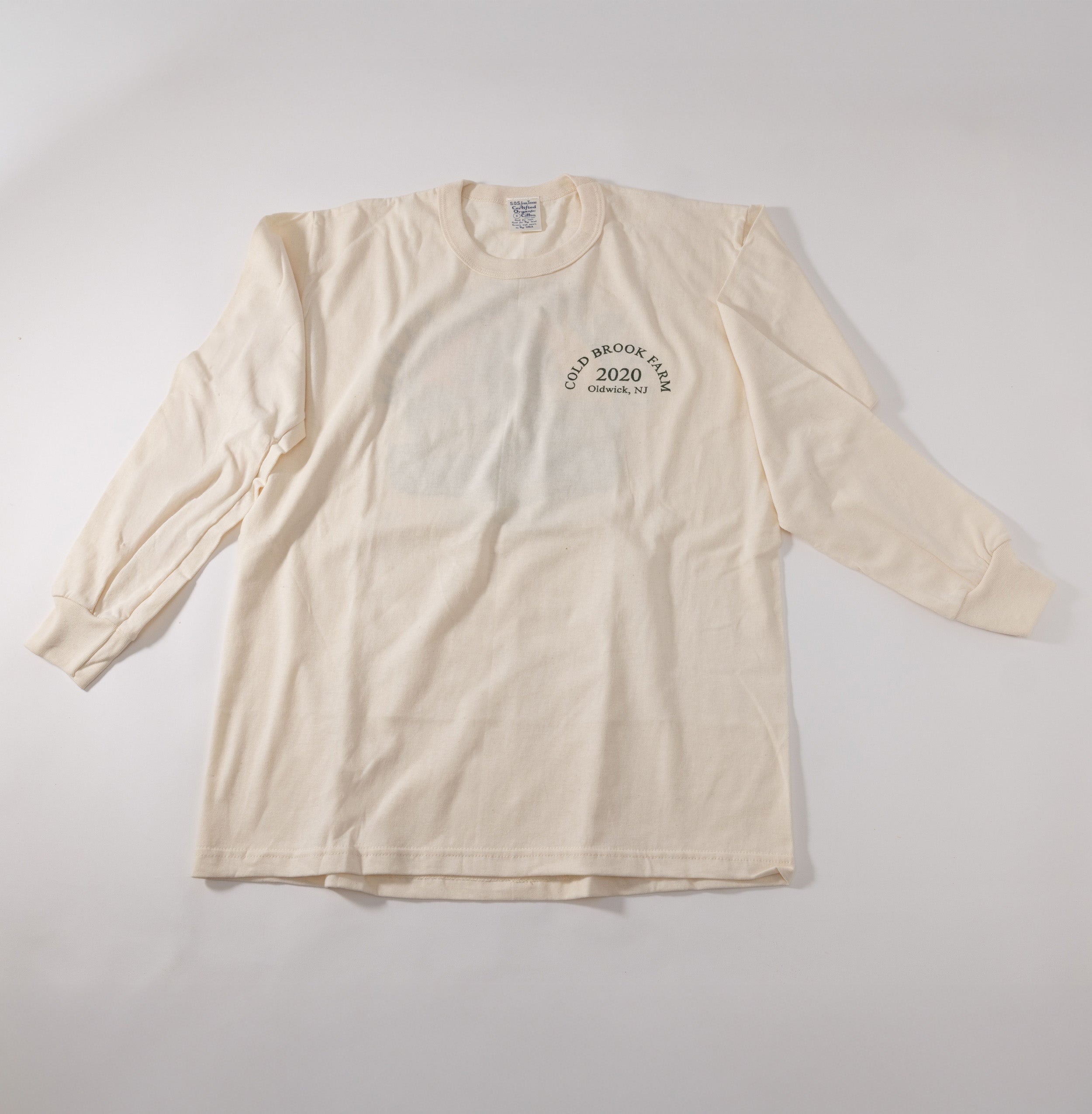A cream-colored long-sleeve t-shirt with a small black circular graphic on the left chest reading Cold Brook Farm 2020 Oldwick, NJ, laid flat on a light gray background.