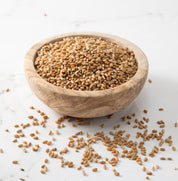 A wooden bowl filled with redeemer whole wheat grains, with a few scattered on a light marble surface.