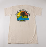An off-white short-sleeve t-shirt laid flat against a light gray background, featuring a large vintage-style graphic on the back with the text 'COLD BROOK FARM', 'Oldwick, NJ', and a colorful farm landscape illustration.