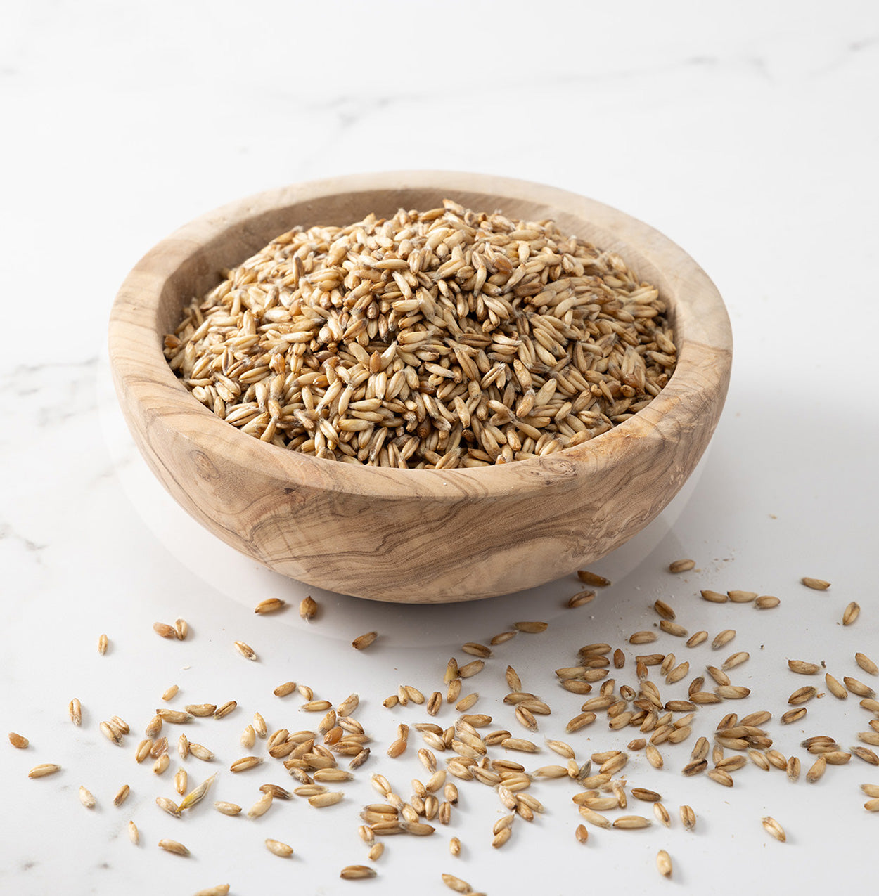A wooden bowl filled with hulless streaker oats, photographed on a light marble-like surface with a few seeds scattered around.