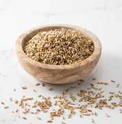 A wooden bowl filled with hulless streaker oats, photographed on a light marble-like surface with a few seeds scattered around.