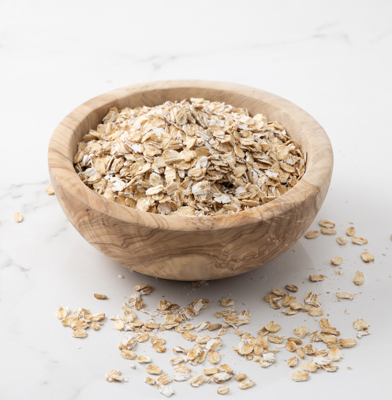 Rolled oats piled in a round wooden bowl on a light marble surface, with a few oats scattered around the bowl.