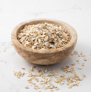 Rolled oats piled in a round wooden bowl on a light marble surface, with a few oats scattered around the bowl.