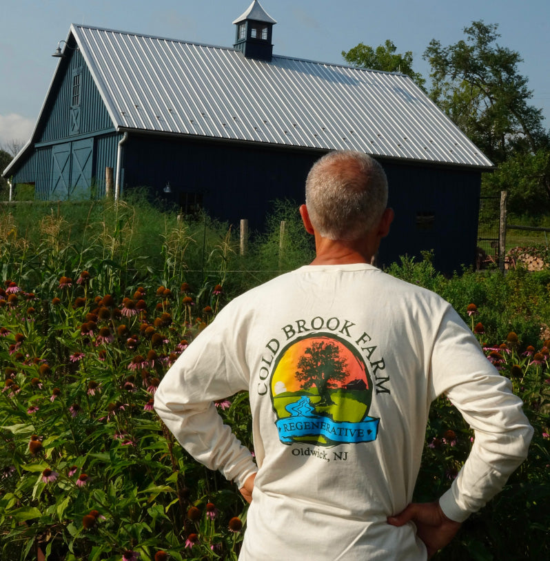 Jason Desalvo viewed from behind wearing a cream long-sleeve t-shirt with a colorful 'Cold Brook Farm' graphic on the back, standing in a garden with a dark blue barn in the background.