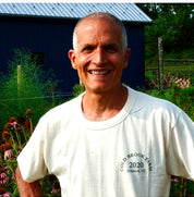 A smiling farmer; Jason Desalvo wearing a white t-shirt with 'COLD BROOK FARM 2020 Oldwick, NJ' printed on the chest, standing outdoors in front of a blue barn and garden plants.