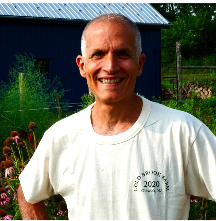 A smiling farmer; Jason Desalvo wearing a white t-shirt with 'COLD BROOK FARM 2020 Oldwick, NJ' printed on the chest, standing outdoors in front of a blue barn and garden plants.