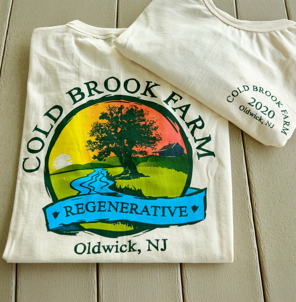 A folded cream-colored t-shirt with a colorful 'Cold Brook Farm Regenerative' graphic, displayed on a light wood plank surface with another folded shirt showing the side print.