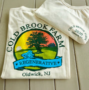 A folded cream-colored t-shirt with a colorful 'Cold Brook Farm Regenerative' graphic, displayed on a light wood plank surface with another folded shirt showing the side print.