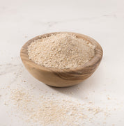 A small round wooden bowl filled with fresh milled oat flour, set on a white marble-like surface with some out flour scattered around it.