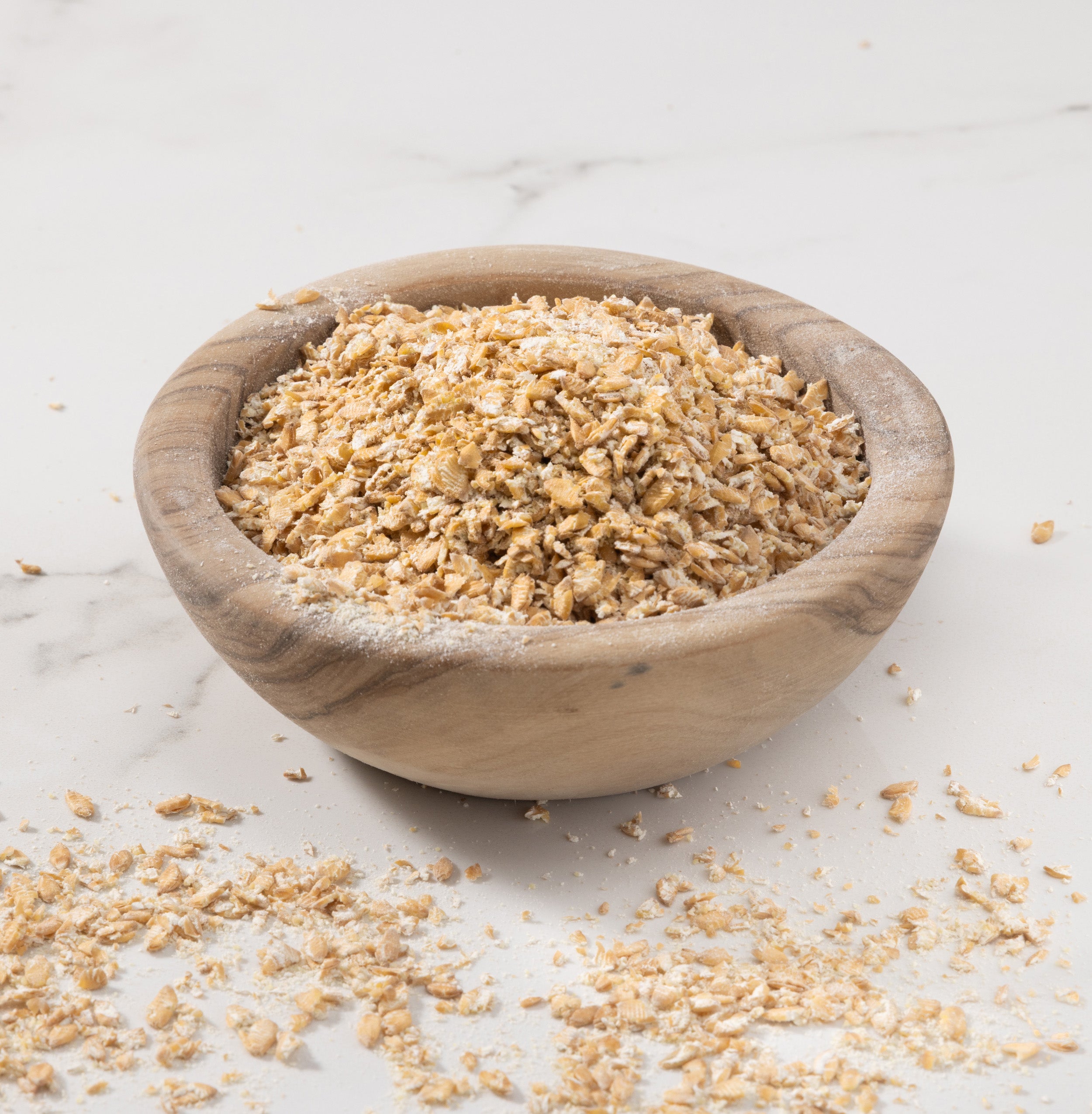 A wooden bowl filled with rolled einkorn, set on a light marble surface with some einkorn flakes scattered around.