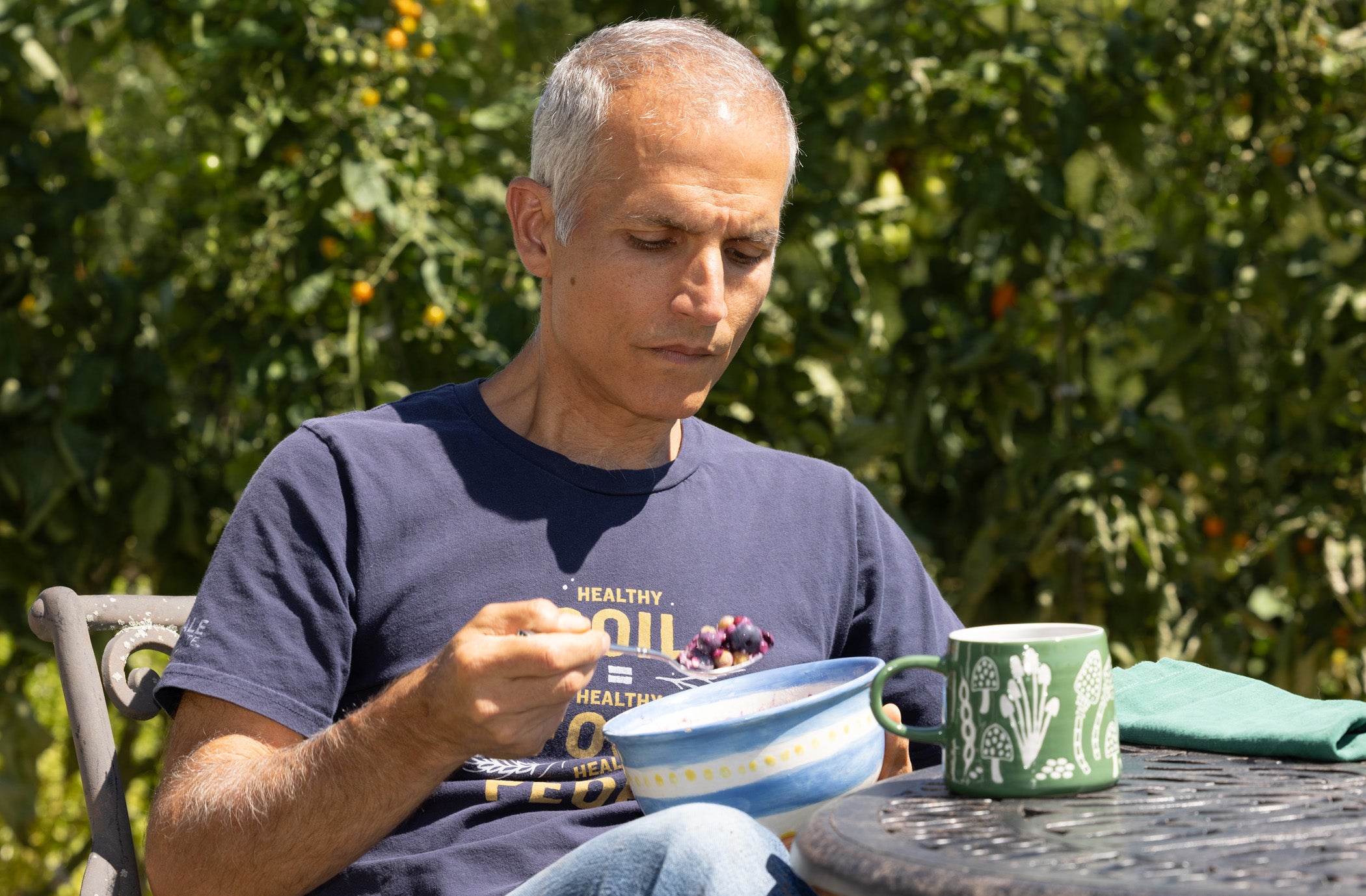 Jason Desalvo sitting outdoors at a table, eating cereal from a blue bowl while wearing a navy blue t-shirt with yellow text. A green mug with white botanical patterns sits on the table next to him, with a green garden background.