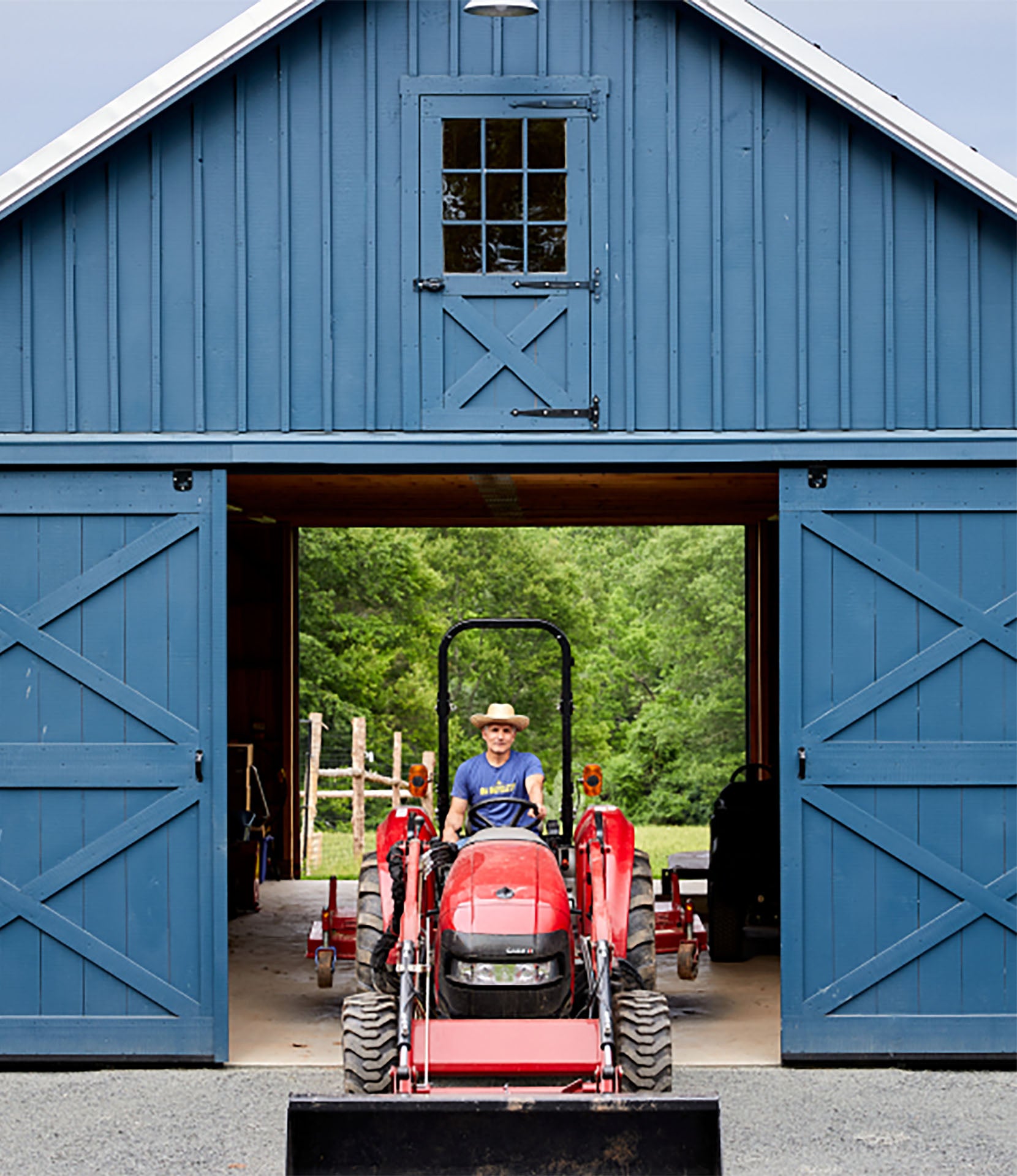 A red tractor with a front loader attachment is parked inside the open doors of a large blue barn, with a person sitting in the tractor. Lush green trees are visible in the background outside the barn.