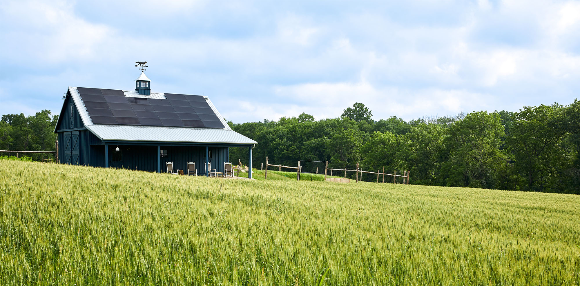 A dark blue barn with solar panels on its roof sits in a green field under a cloudy blue sky, with a line of trees in the background.