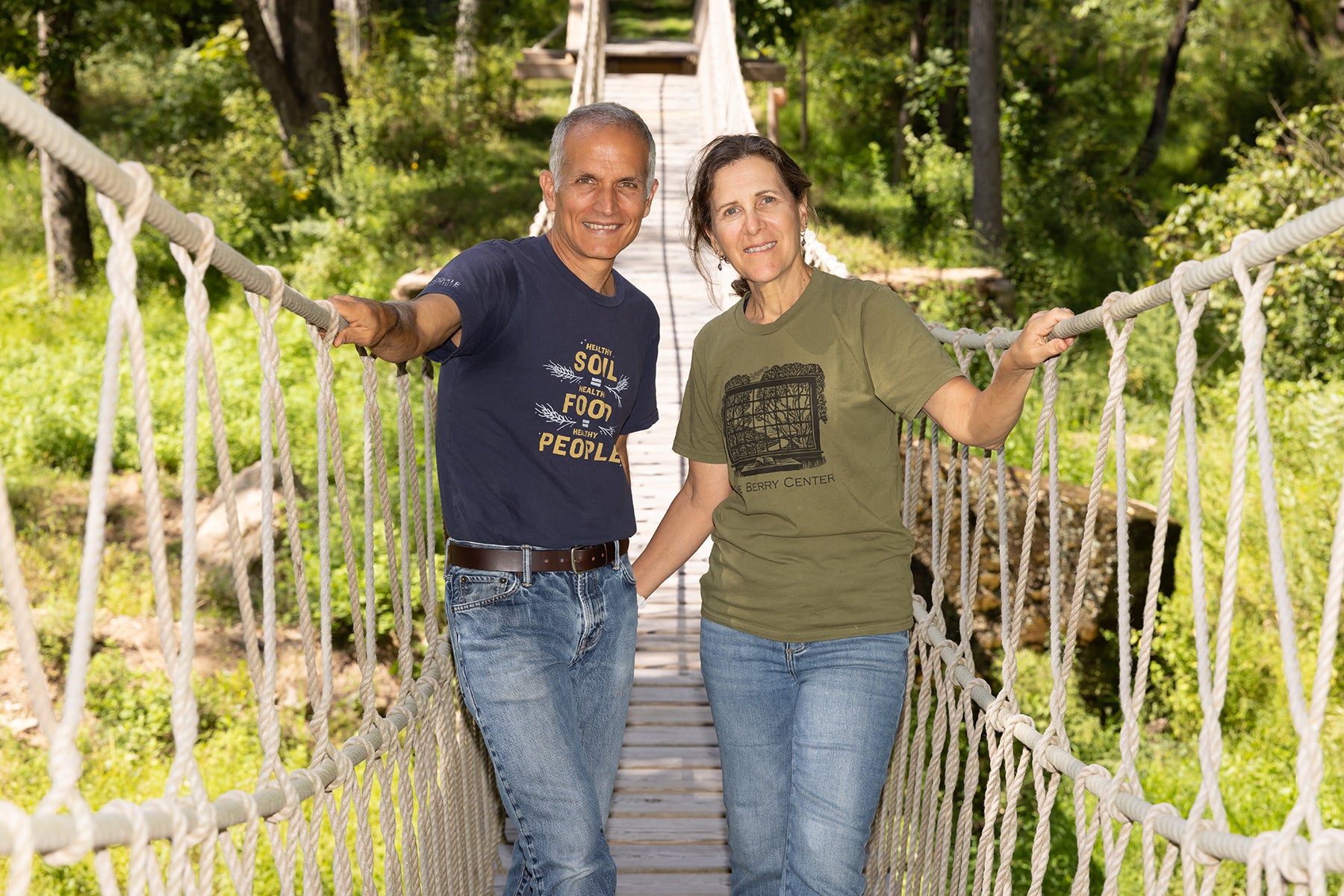 A man and a woman standing together on a rope suspension bridge in a lush green outdoor setting. The man wears a navy blue graphic t-shirt and jeans, while the woman wears an olive green graphic t-shirt and jeans.