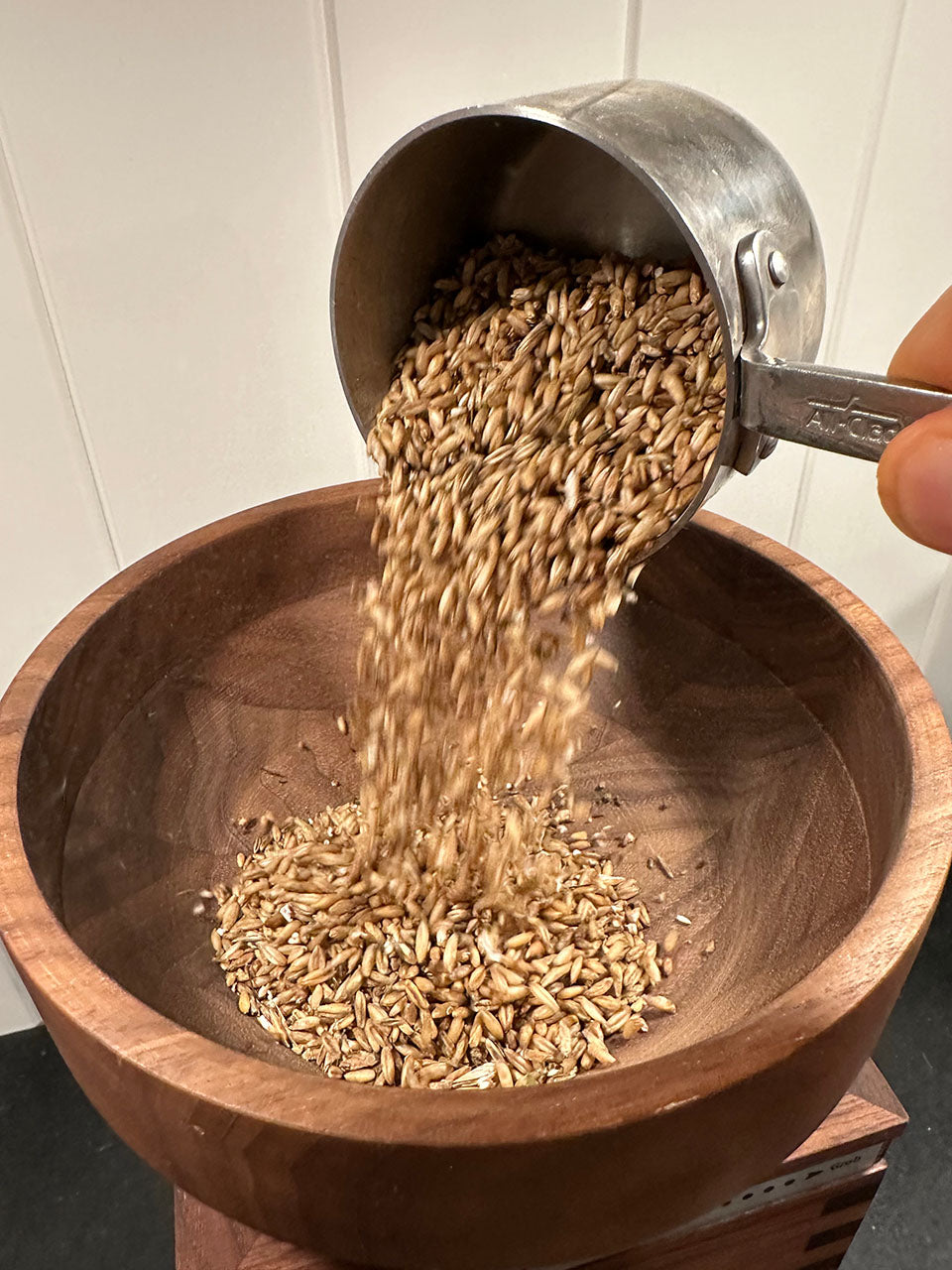 A metal measuring cup pouring brown grains into a large wooden bowl, with a hand holding the cup against a light tiled background.