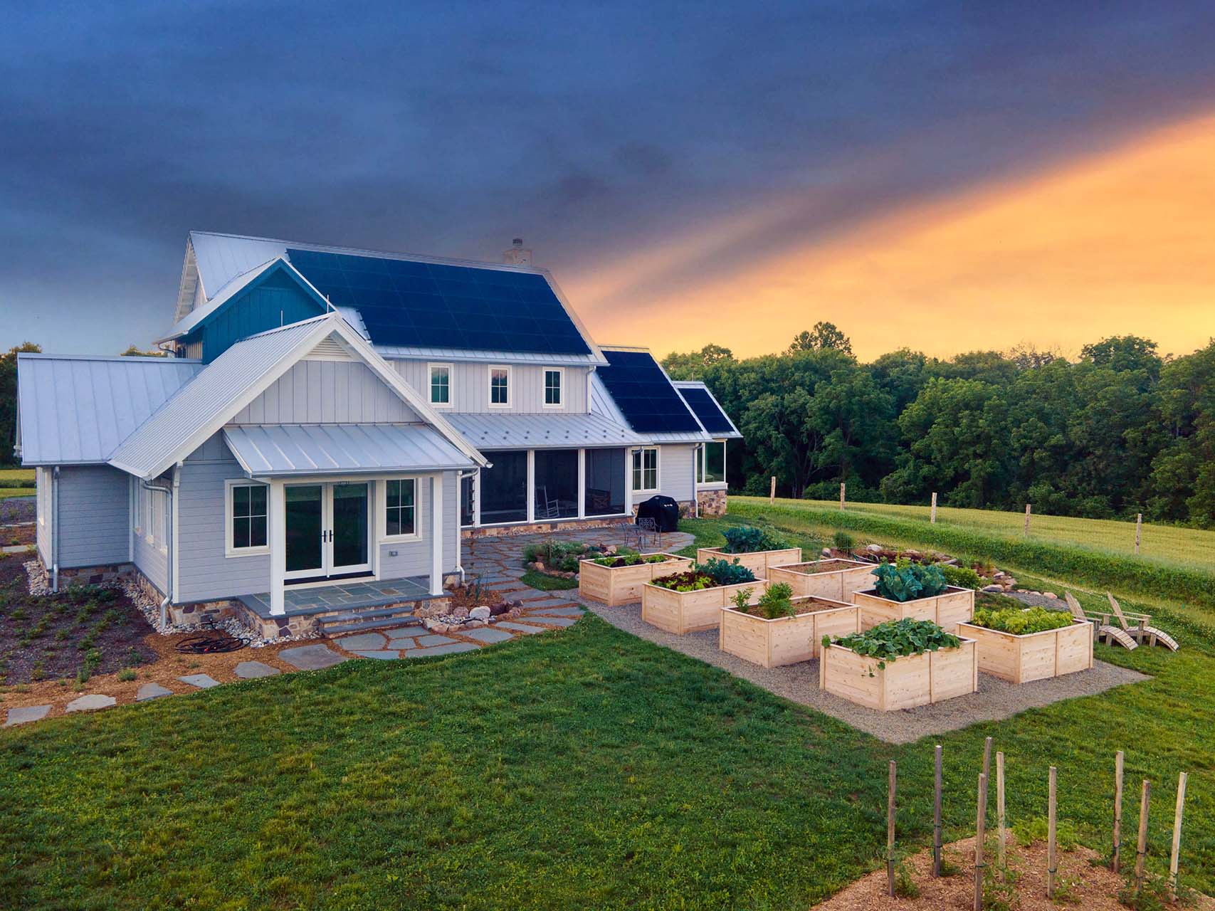 A large white farmhouse-style home with dark metal roofing and solar panels, shown from an elevated outdoor perspective. The house is set on a grassy lawn with raised wooden garden beds, trees in the background, and a dramatic sunset sky with blue clouds and orange light.